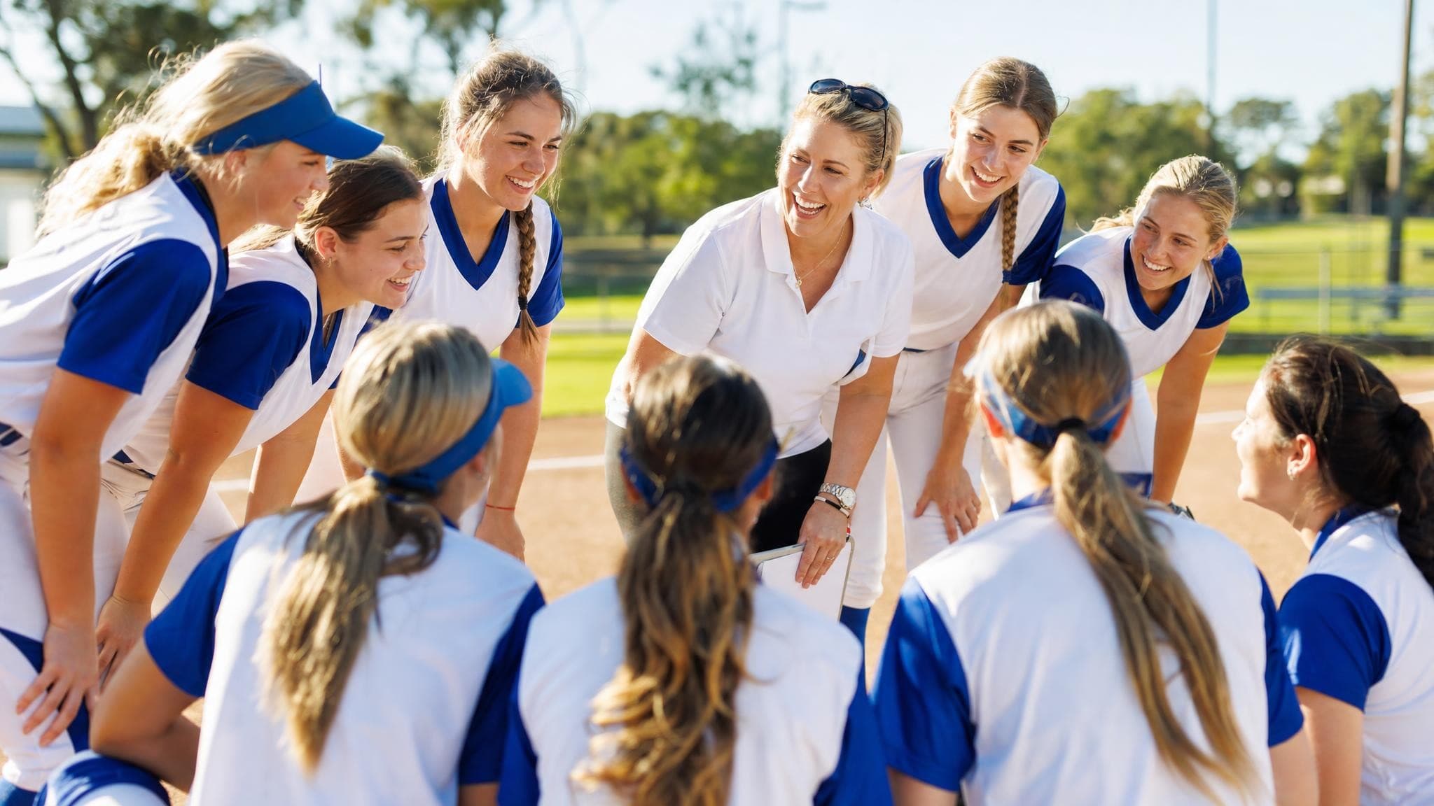 A girls’ softball team in blue and white huddled around their laughing coach, who holds a clipboard, on a sunlit infield.