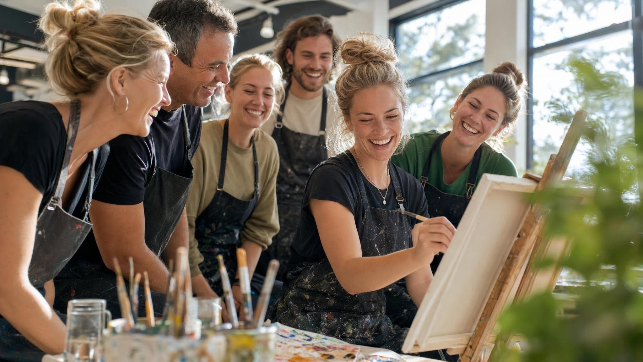 A small group of adults in paint-spattered aprons laughing around a tutor painting at an easel in a bright studio.