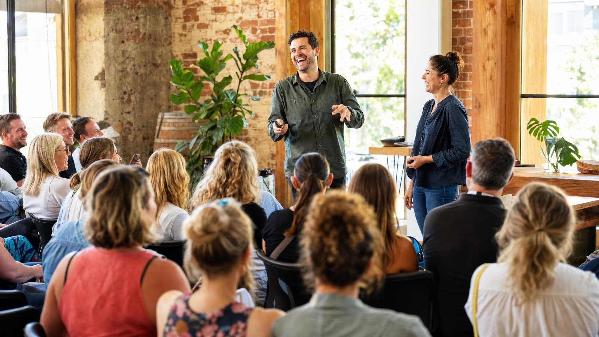 A packed audience in a brick-walled workshop space laughing along with two hosts presenting at the front.
