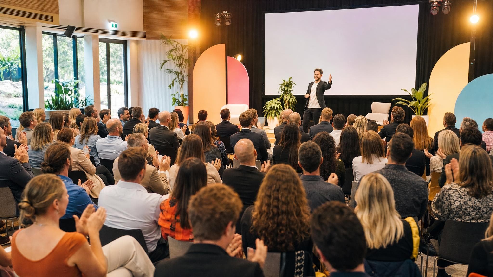 A packed conference hall of adults laughing and applauding a speaker on a bright stage.