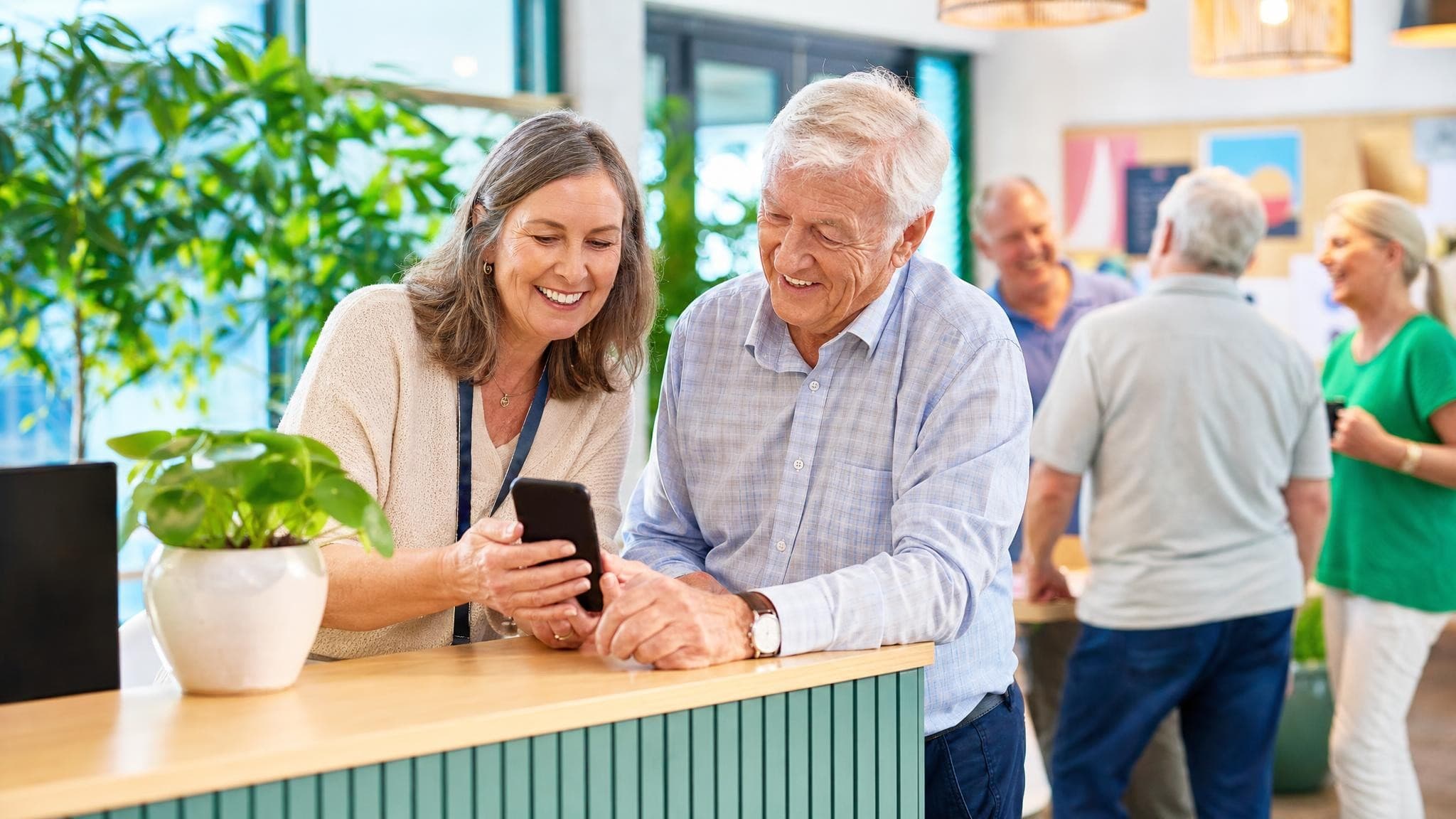 A smiling community-centre coordinator at the welcome desk showing an older man how to join the centre’s app on his phone, visitors chatting behind them.