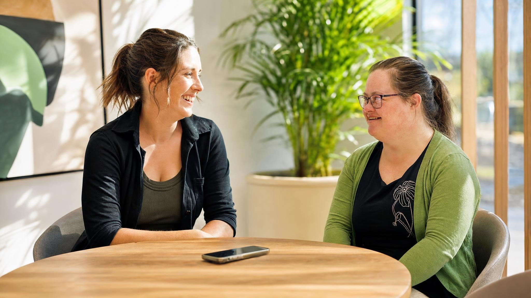 A support worker and a woman in a green cardigan smiling at each other across a round table in a sunlit room, a phone on the table between them.