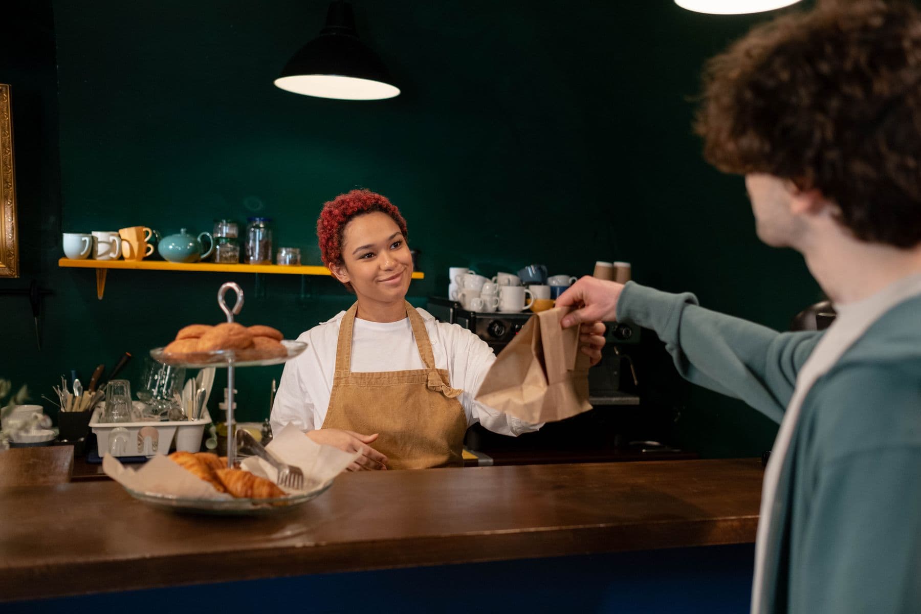 A barista in an apron passing a paper bag of pastries across a café counter to a customer reaching for it.