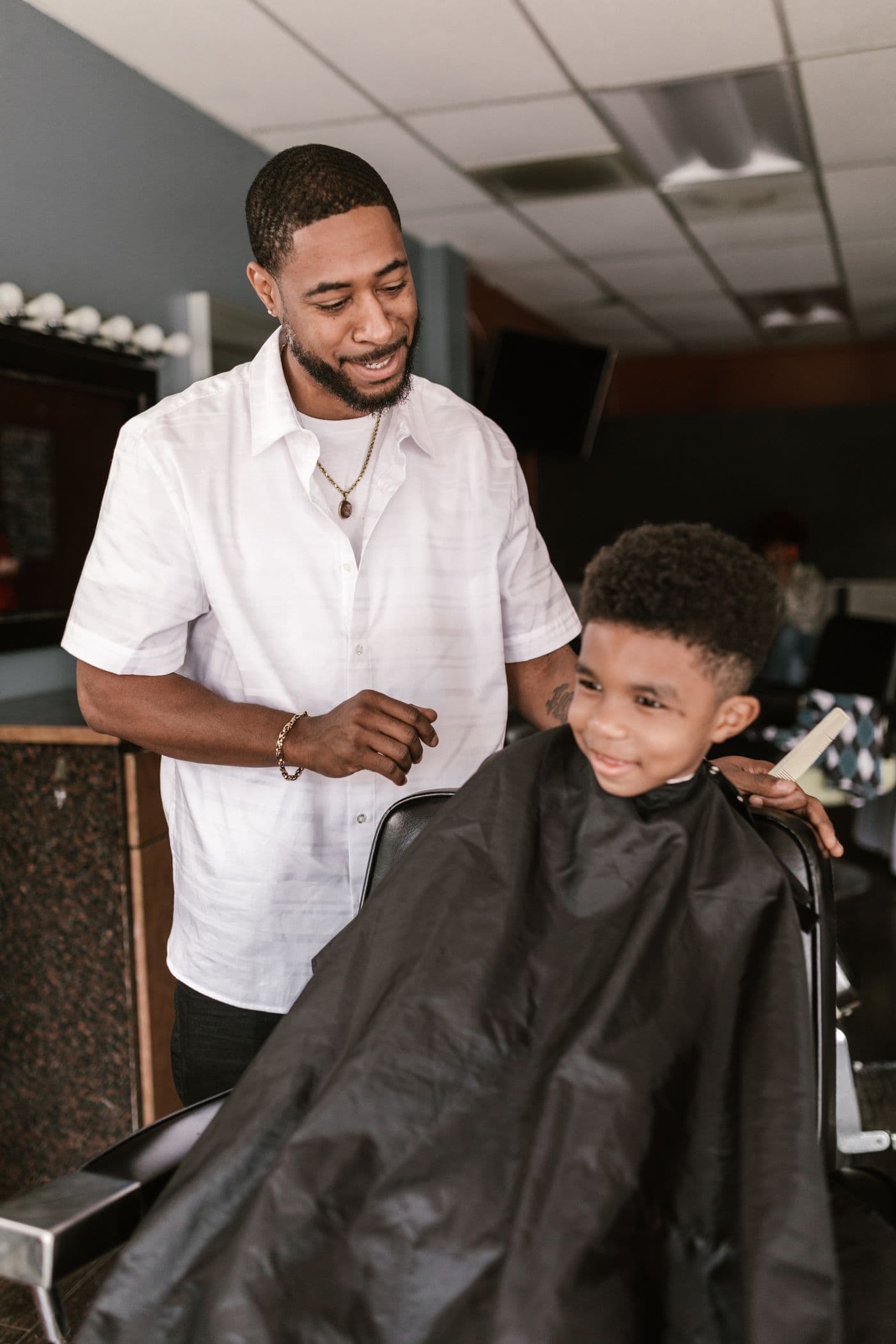 A barber in a white shirt mid-cut, talking and laughing with a young boy sitting in the chair in a cape.