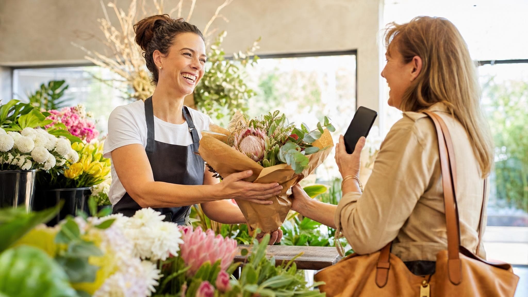 A smiling florist hands a bouquet across her counter to a customer who is tapping her phone to pay.