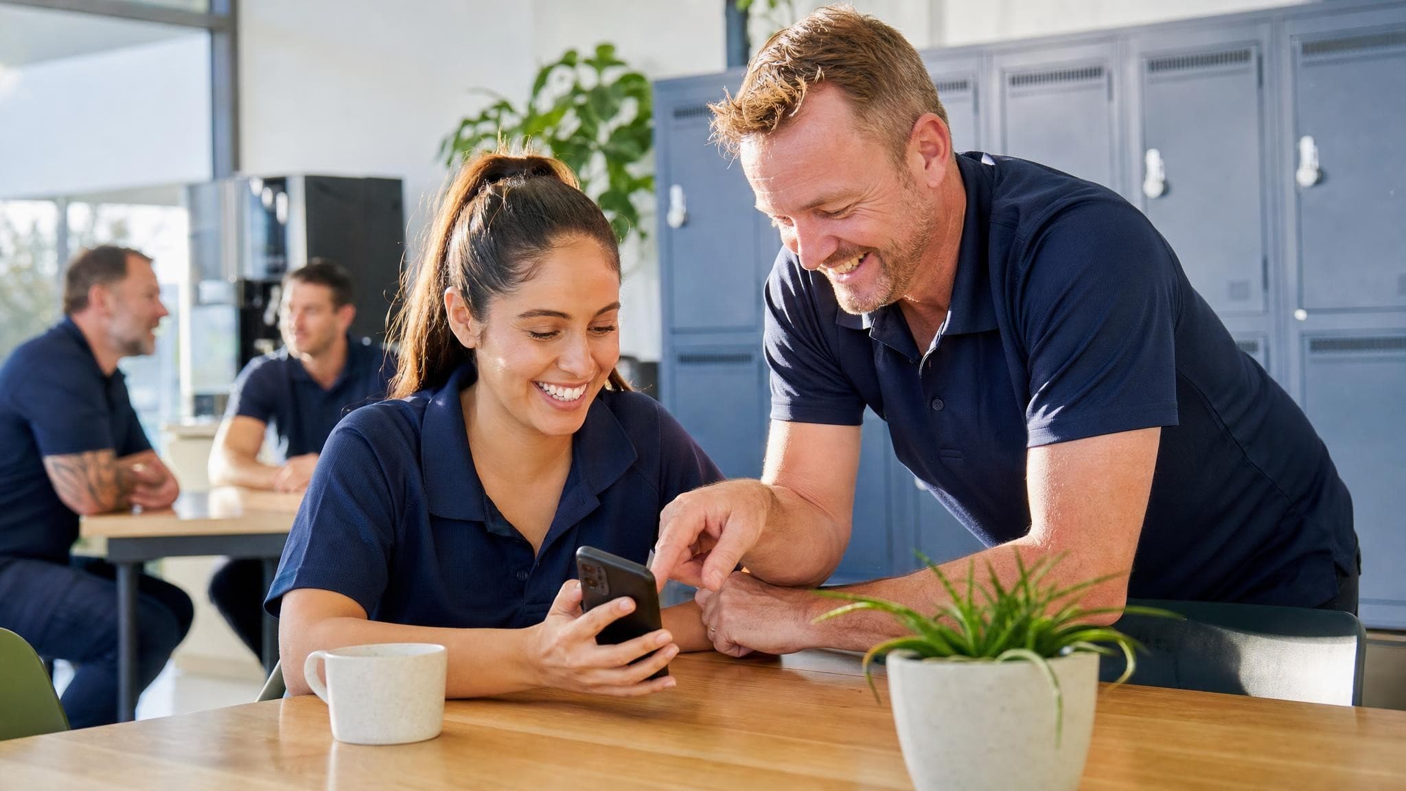 A new staff member in a branded polo reading on her phone in a bright staff room while a colleague points something out and smiles.
