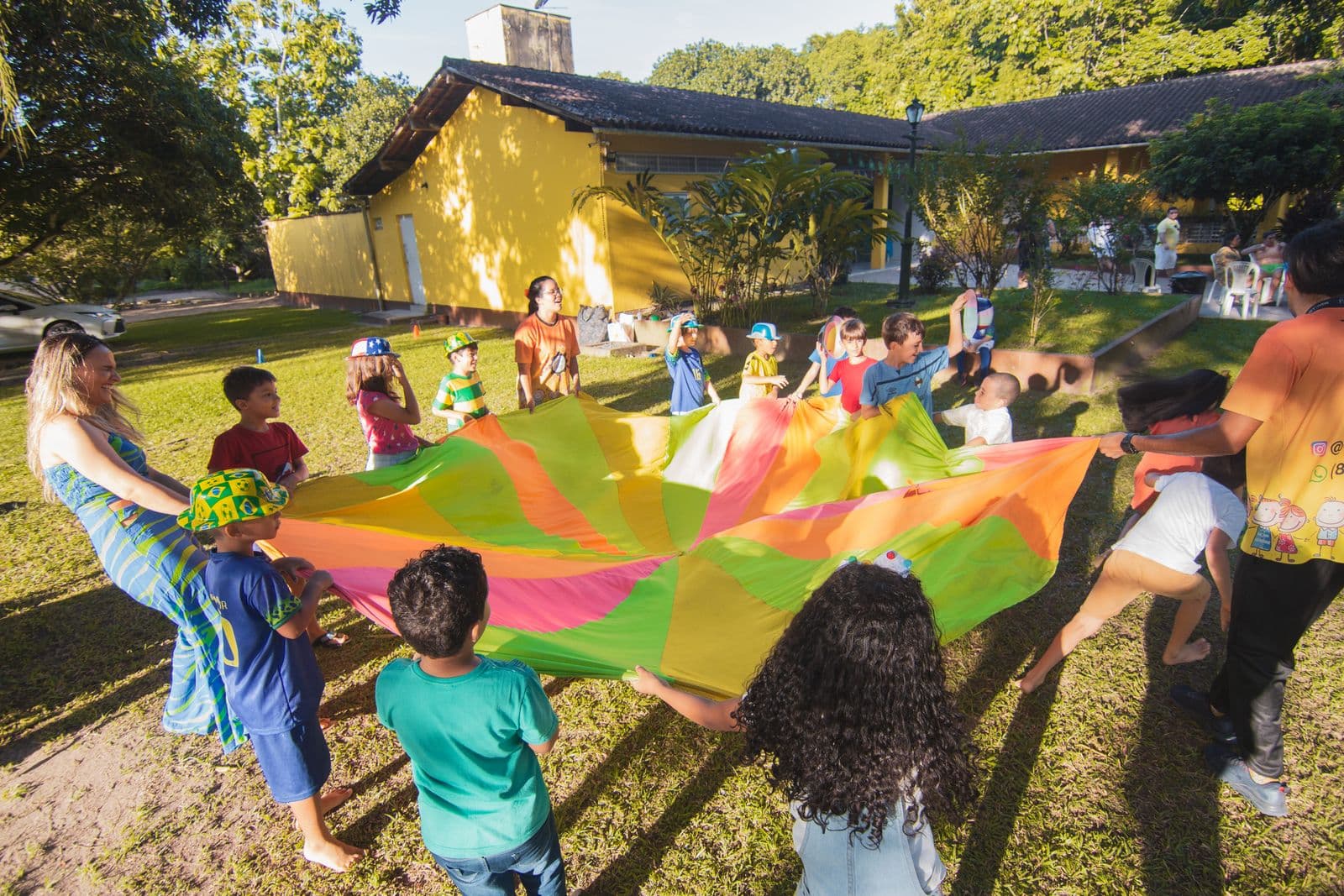 Children and adults outdoors holding the edges of a large colourful parachute at a community gathering in late afternoon sun.