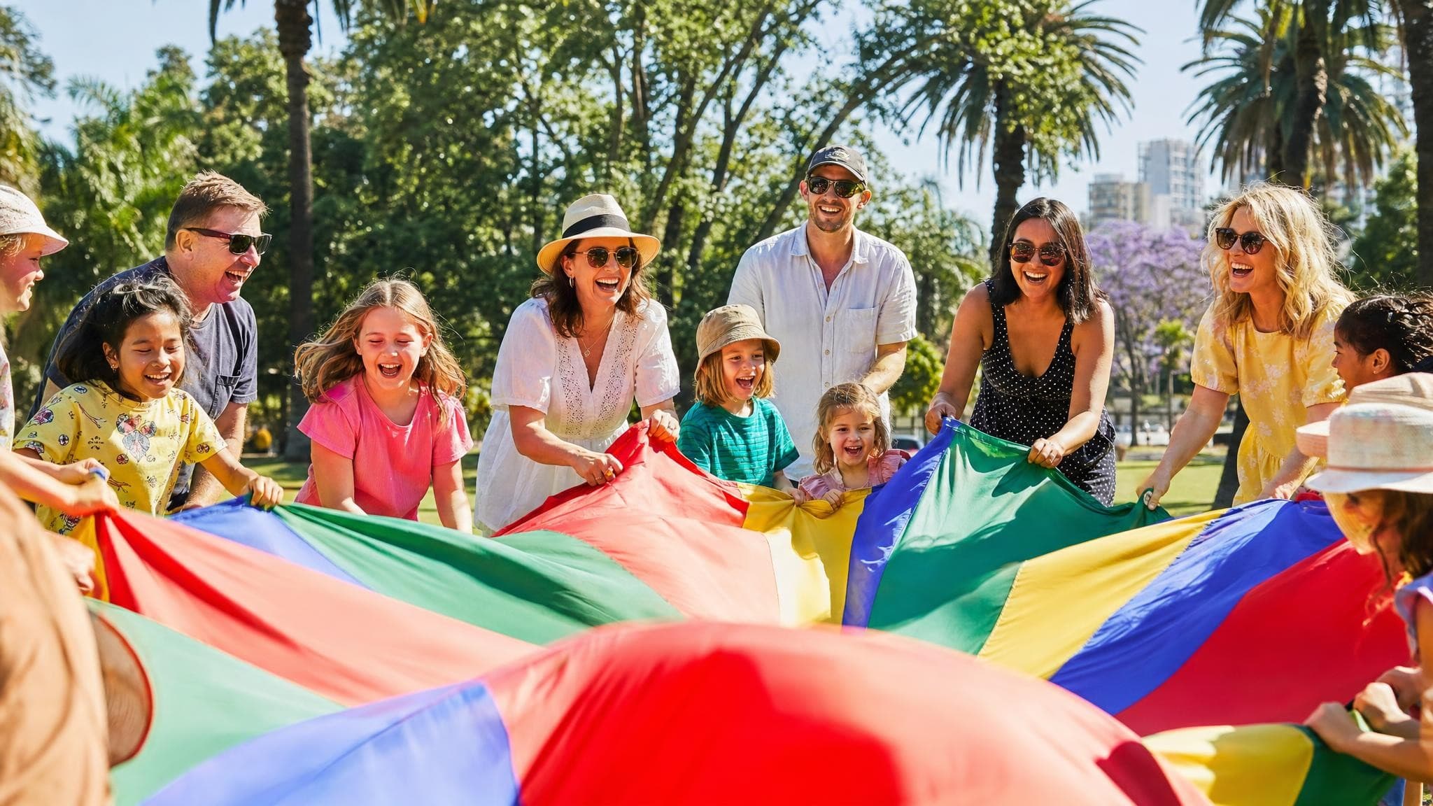Children and adults laughing as they hold the edges of a giant rainbow parachute at a sunny community day in a park.