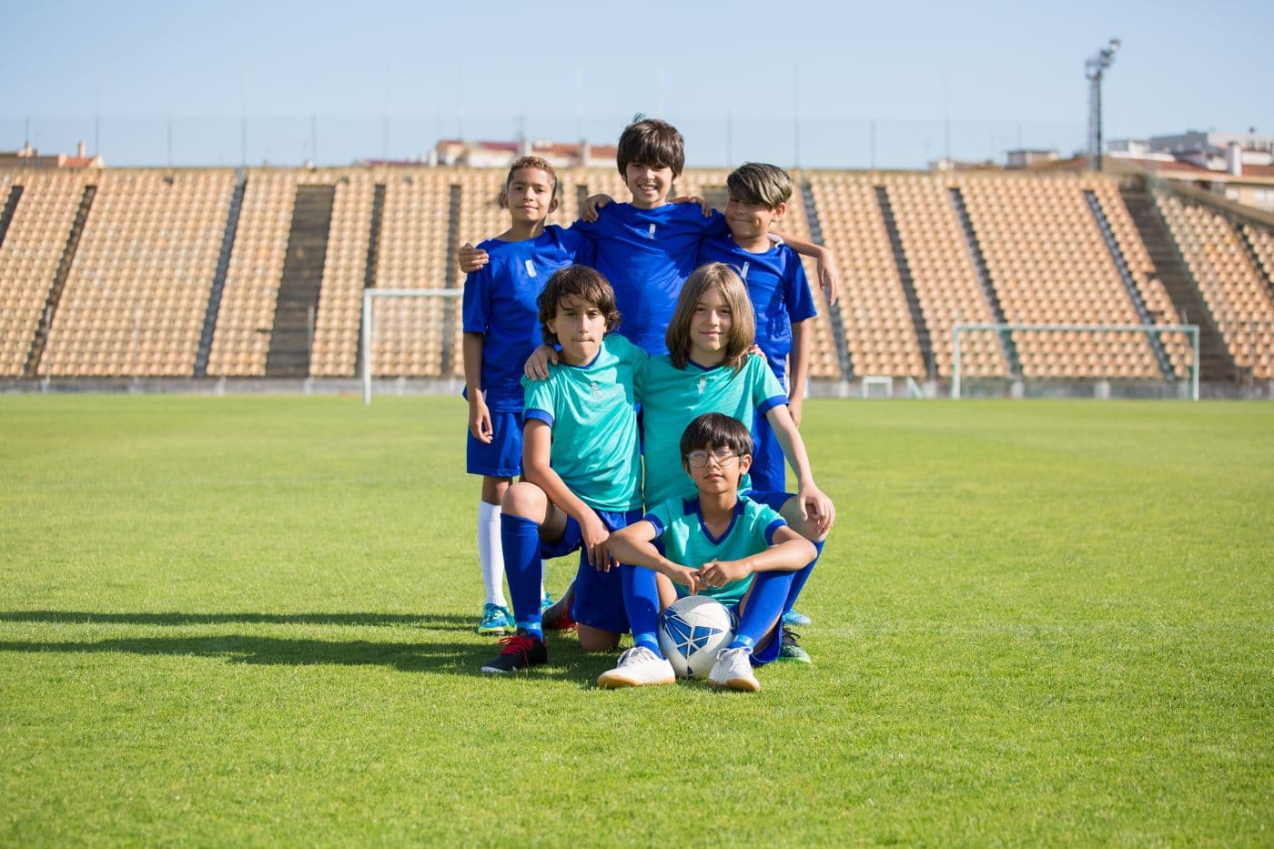 A junior football team in blue and teal kit posed together on a grass pitch with a ball, an empty stand behind them.