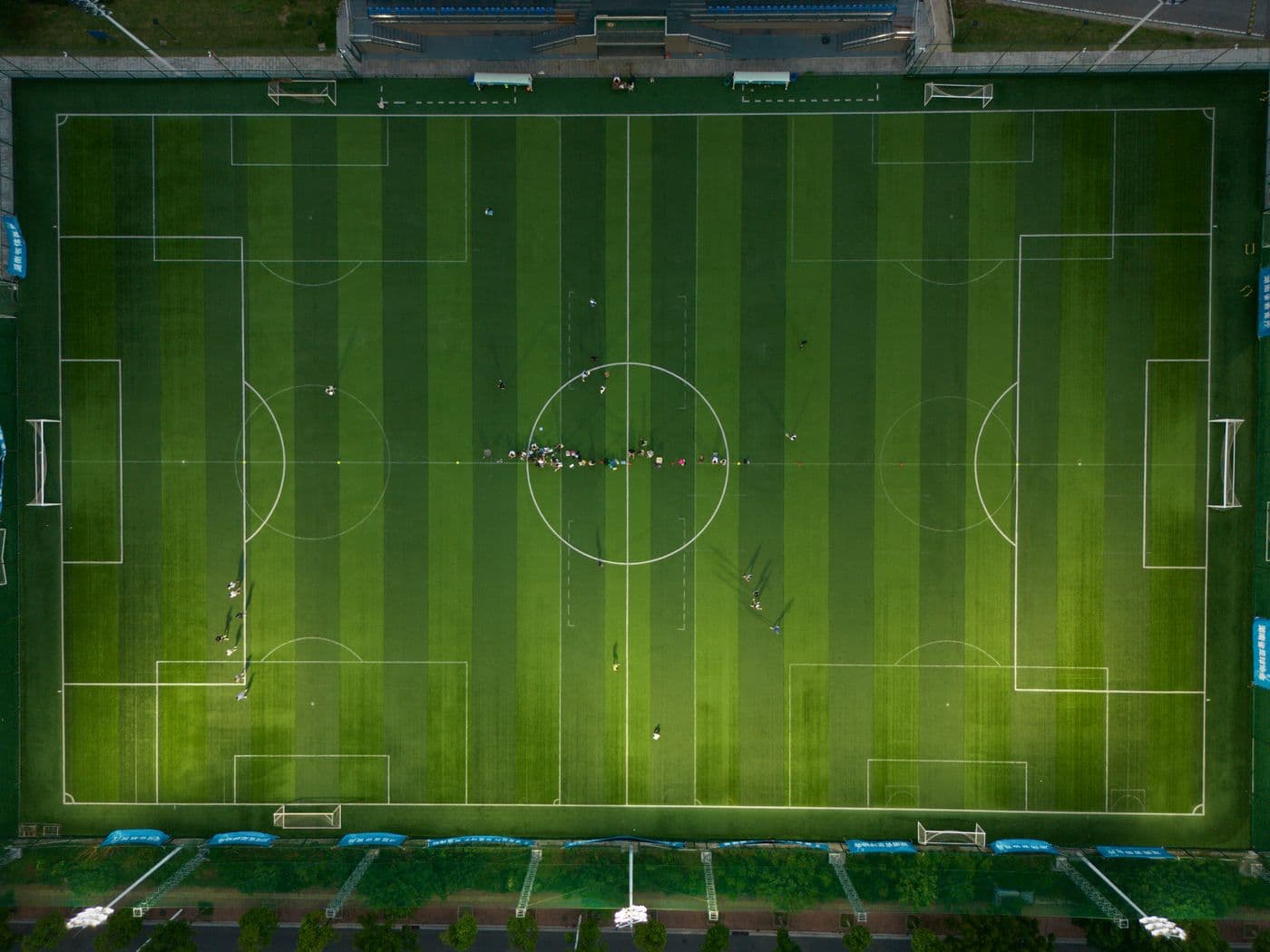 An aerial view of a full-size football pitch that is almost entirely empty, with one small cluster of people gathered near the centre circle.
