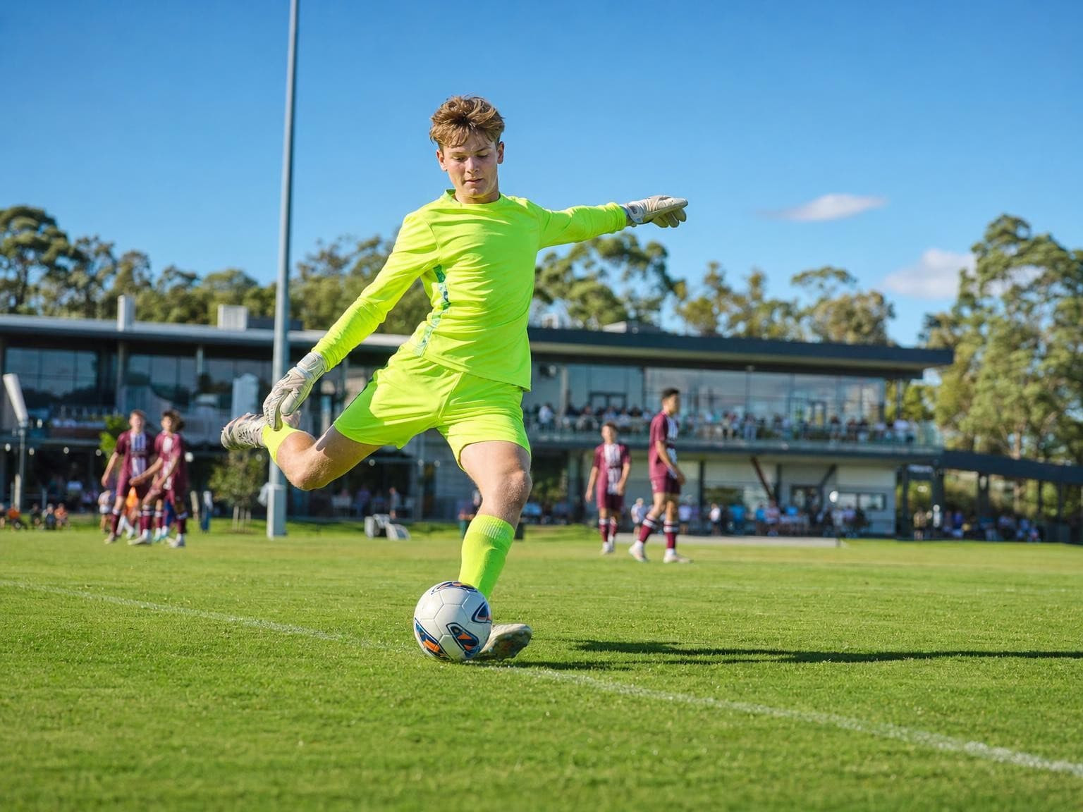 A goalkeeper in a fluorescent yellow kit about to kick the ball upfield, a clubhouse and other players behind him.