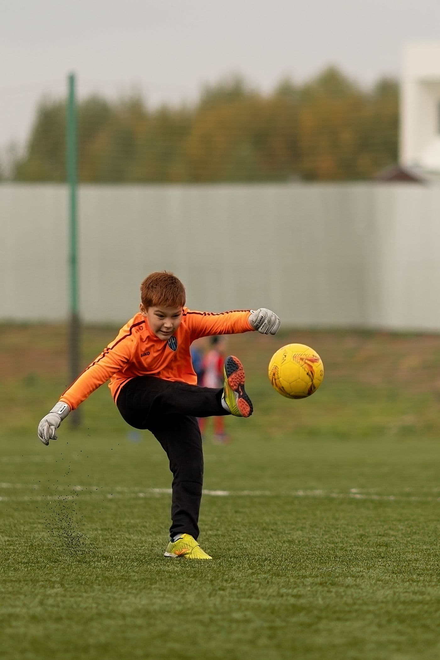 A young goalkeeper in an orange kit kicking a ball on a grass pitch on an overcast day, with a suburban ground behind.