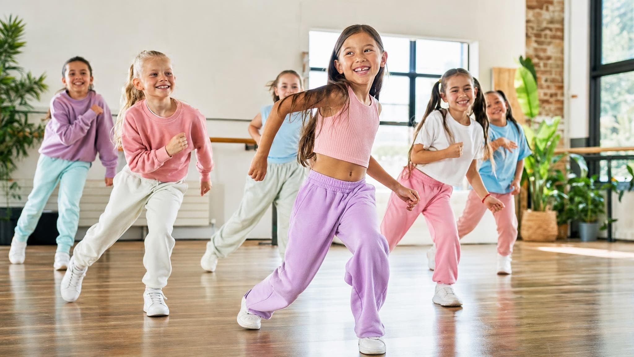 A children’s dance class in a bright studio, a girl mid-step in front and the others following behind her, all smiling.